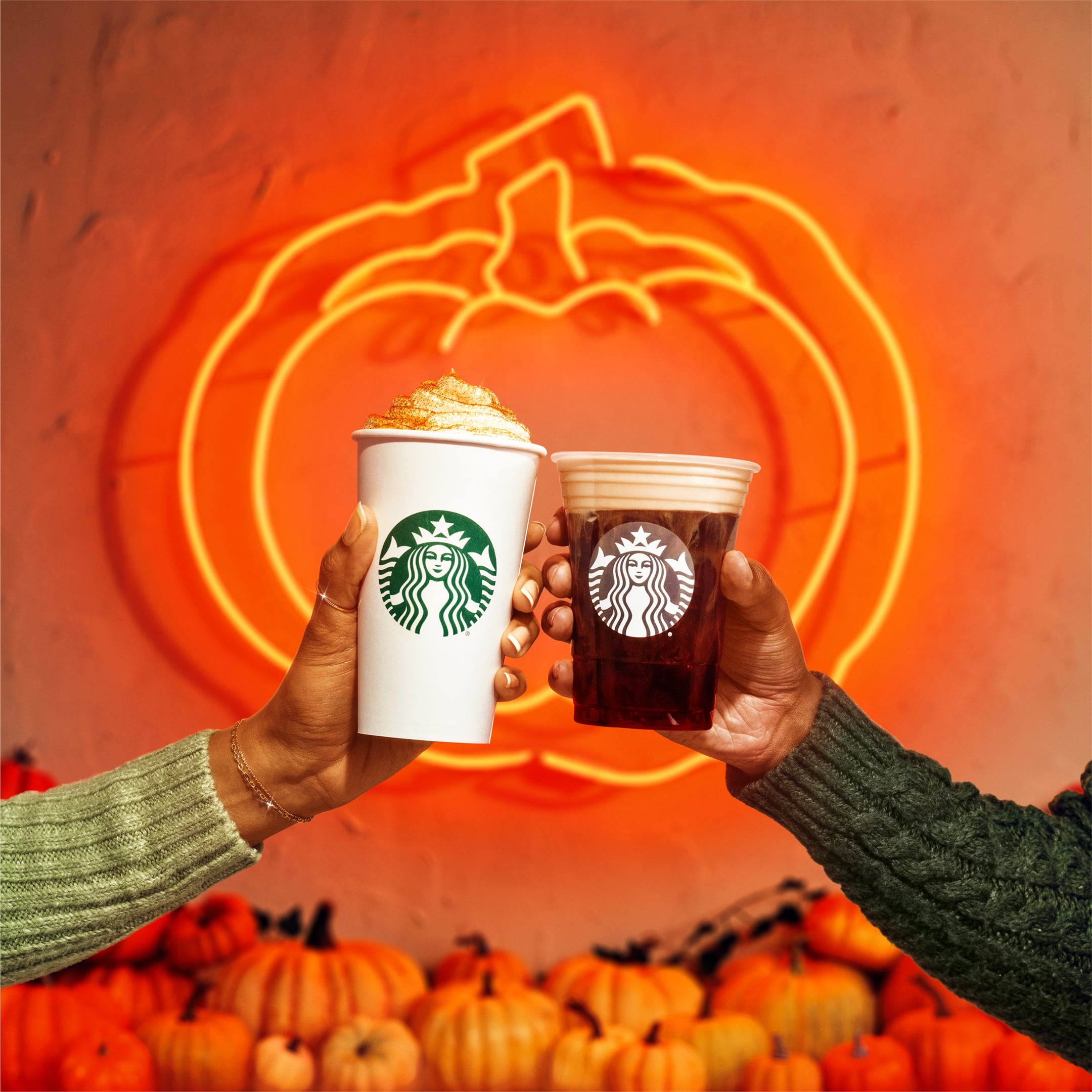 Woman's hand with hot PSL next to man's hand with an iced Pumpkin Spice Cold Brew, in front of orange, pumpkin-themed background