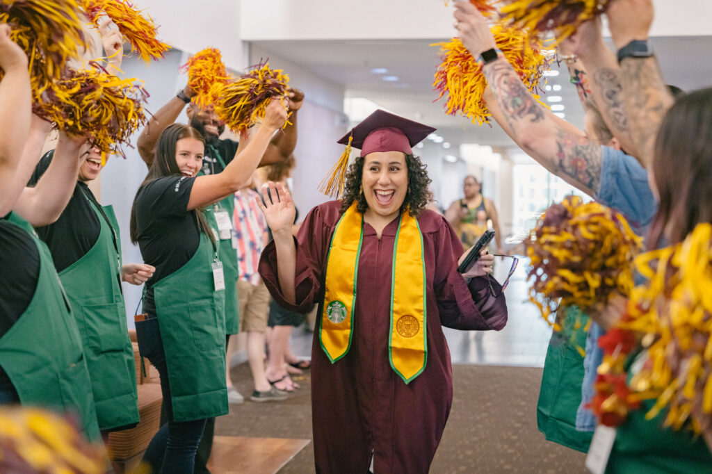 An ASU graduate in cap and gown walking down a row with Starbucks baristas waving pom-poms