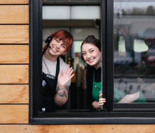 Two female Starbucks baristas wave and smile at an open drive thru window.