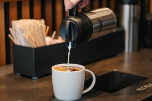 A white mug with coffee and a person pouring milk in the cup on the condiment bar