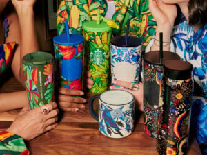 An assortment of FARM Rio and Starbucks collaboration reusable cups on a table and hands holding some of the cups