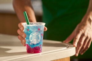 A hand of a Starbucks barista handing over Starbucks Summer Berry Refresher placed on the white counter top