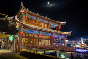A brightly lit traditional building with ornate architecture, featuring colorful decorations and red lanterns, set against a night sky with a visible moon.