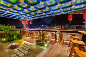 A vibrant rooftop bar features colorful ceiling designs, wooden stools, and lanterns, overlooking a scenic night view.