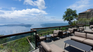 A scenic outdoor seating area features modern lounge chairs and tables, overlooking a vast lake and lush green hills under a blue sky.