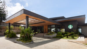 A modern building entrance features a wooden overhang, large glass doors, and lush greenery surrounding stone steps.