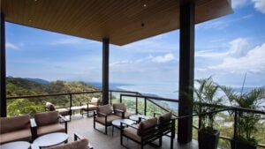 A spacious outdoor terrace features comfortable seating with beige cushions and wooden frames, overlooking a lush green landscape and a body of water under a blue sky.