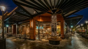 A modern café with a wooden exterior and large glass windows. Inside, there are stylish seating areas and a decorative column featuring intricate designs. Outdoor seating is arranged with small tables and woven chairs. The space is well-lit, creating a warm ambiance.