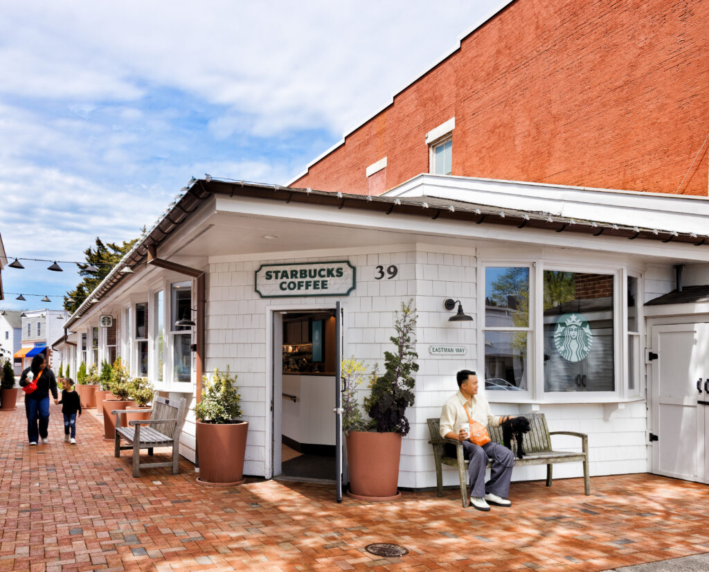 A Starbucks coffee shop features a white exterior with a green sign. A man sits on a bench outside, holding a drink and a small dog. The area has potted plants and a brick walkway.