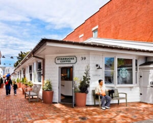 A Starbucks coffee shop features a white exterior with a green sign. A man sits on a bench outside, holding a drink and a small dog. The area has potted plants and a brick walkway.