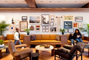 A cozy lounge area features a brown leather sofa against a gallery wall of framed art and photographs. A round marble coffee table sits in front, surrounded by various seating options. Green plants add a touch of nature to the space.