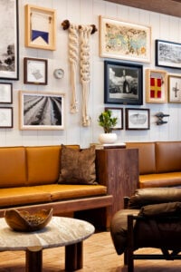 A cozy seating area features a brown leather sofa with a textured cushion, a round marble coffee table, and a wooden side table. The wall is adorned with various framed artworks, including photographs and decorative items, alongside a macramé piece. A small potted plant adds a touch of greenery.