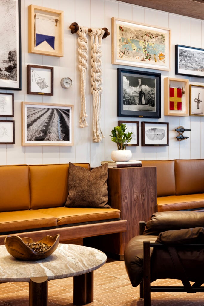 A cozy seating area features a brown leather sofa with a textured cushion, a round marble coffee table, and a wooden side table. The wall is adorned with various framed artworks, including photographs and decorative items, alongside a macramé piece. A small potted plant adds a touch of greenery.