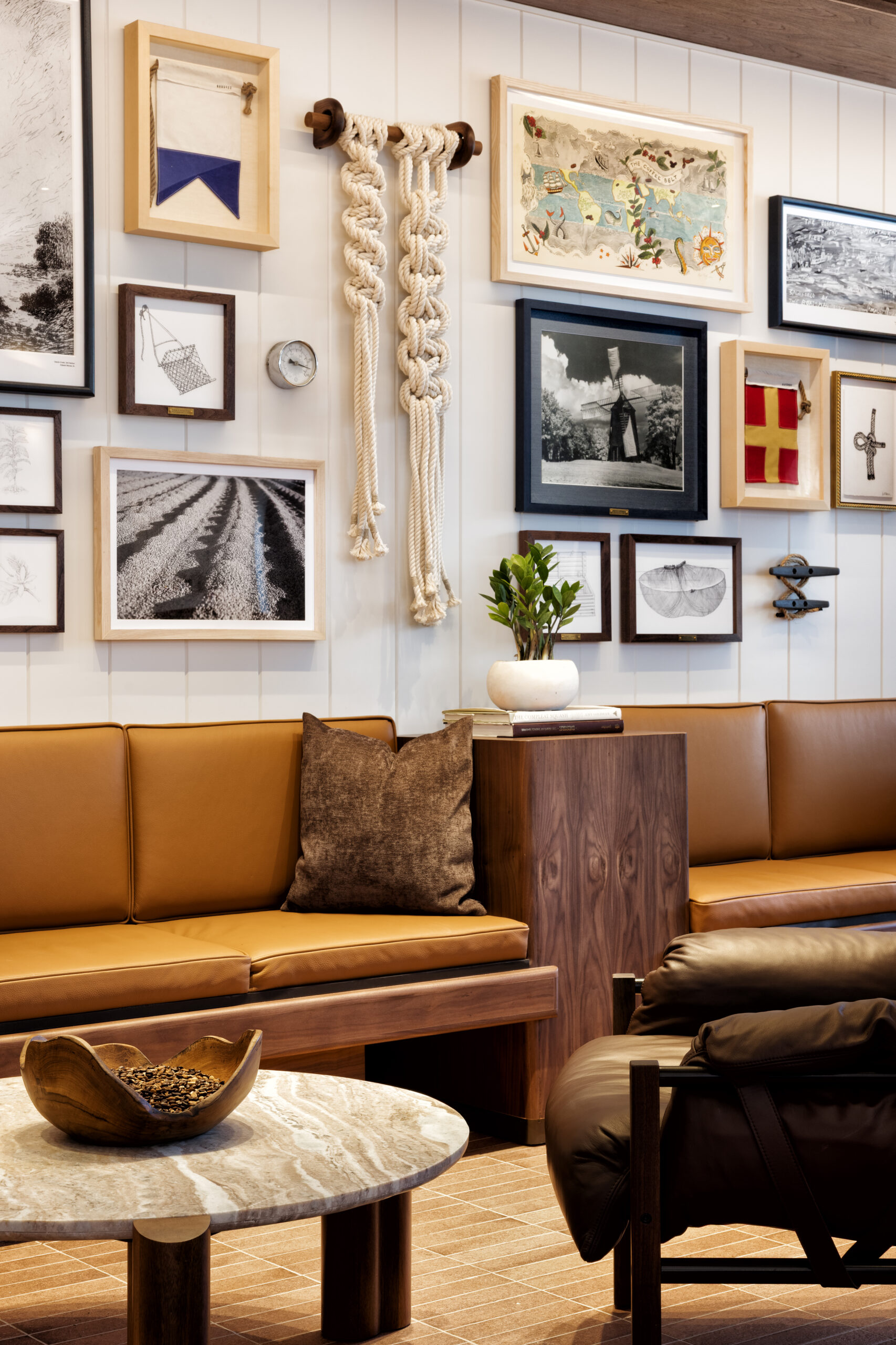 A cozy seating area features a brown leather sofa with a textured cushion, a round marble coffee table, and a wooden side table. The wall is adorned with various framed artworks, including photographs and decorative items, alongside a macramé piece. A small potted plant adds a touch of greenery.
