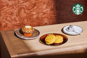 A wooden table displays a pumpkin cream cheese muffin on a gray plate, alongside two egg bites on an amber plate, and a raccoon cake pop on a white plate.