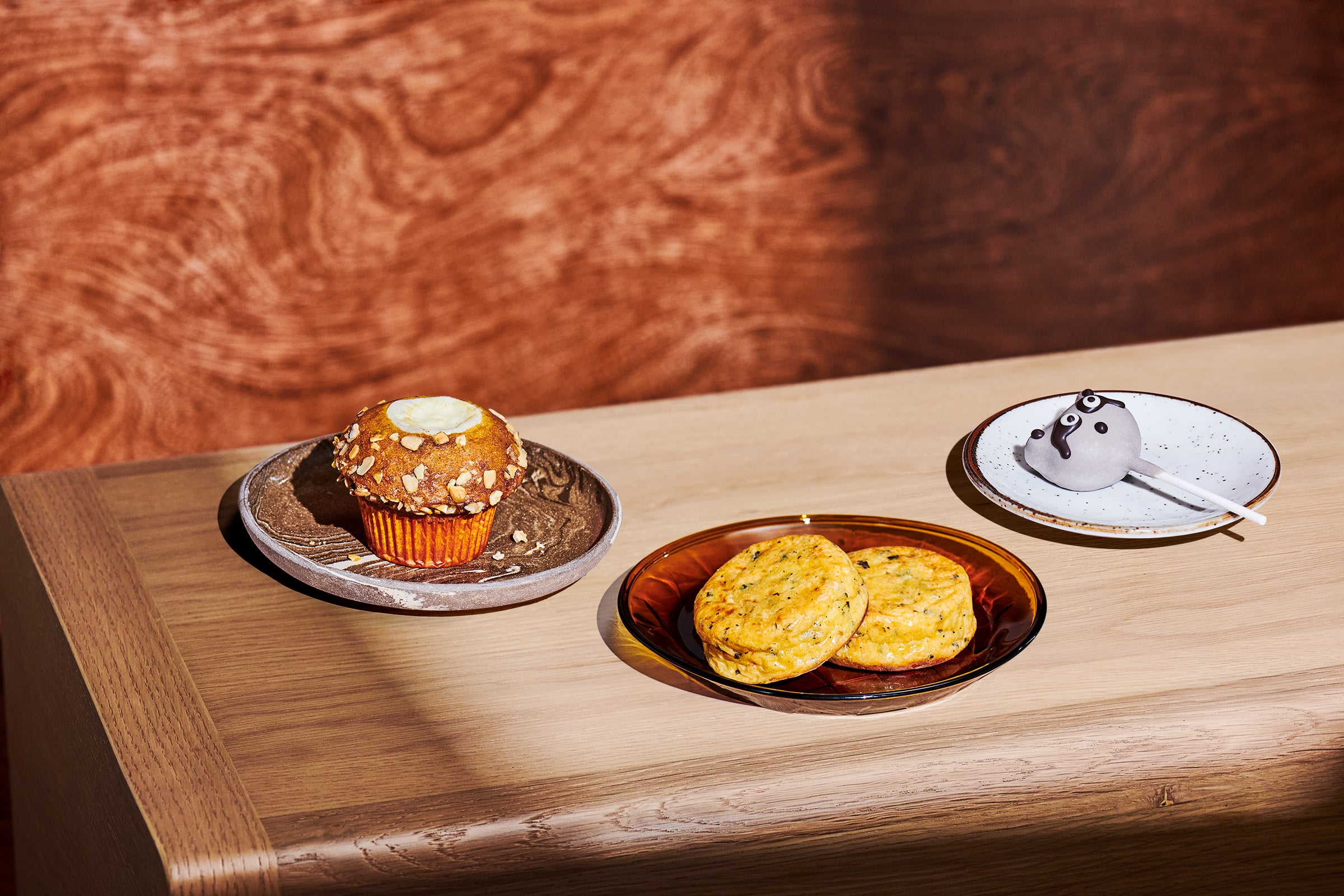 A wooden table displays a muffin topped with oats on a gray plate, alongside two egg bites on a brown glass plate, and a racoon cake pop on a white plate.
