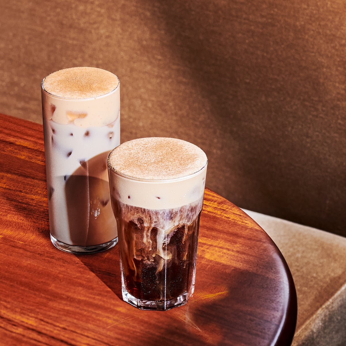 Two glasses of iced coffee drinks are placed on a wooden table. The taller glass contains a creamy, light-colored beverage with ice, while the shorter glass has a darker coffee with a frothy top. Both drinks have a dusting of cinnamon or similar topping.