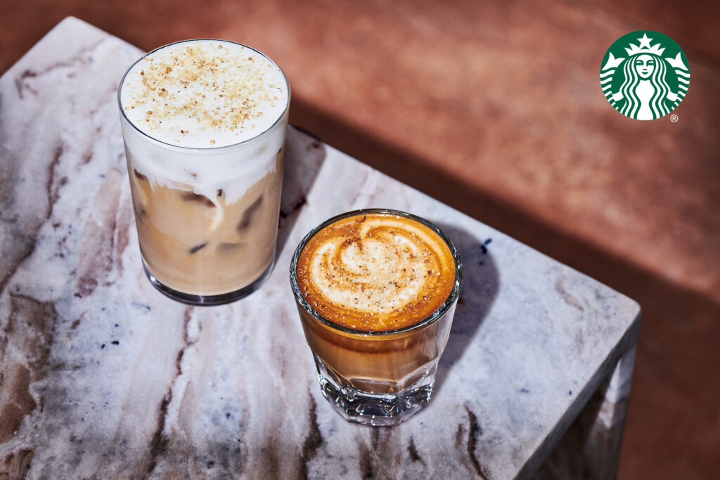 Two coffee drinks are displayed on a marble surface. On the left, a tall glass of iced coffee topped with frothy cream and sprinkled with sugar. On the right, a short glass of espresso with a creamy swirl and a dusting of spice on top.