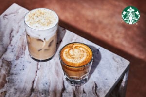 Two coffee drinks are displayed on a marble surface. On the left, a tall glass of iced coffee topped with frothy cream and sprinkled with sugar. On the right, a short glass of espresso with a creamy swirl and a dusting of spice on top.