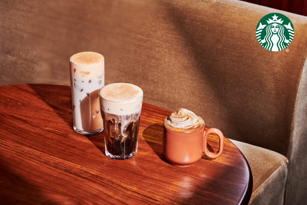 Three coffee drinks are displayed on a wooden table: a tall glass of iced coffee with a frothy top, a shorter glass of dark coffee with foam, and a small orange mug topped with whipped cream and a sprinkle of cinnamon.