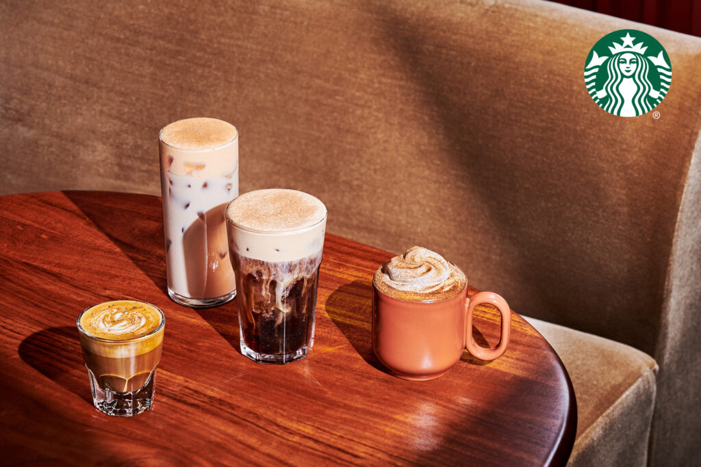 A wooden table displays four coffee drinks: a tall glass of iced coffee, a shorter glass with frothy coffee, a small espresso, and a mug with whipped cream and a sprinkle of cinnamon.