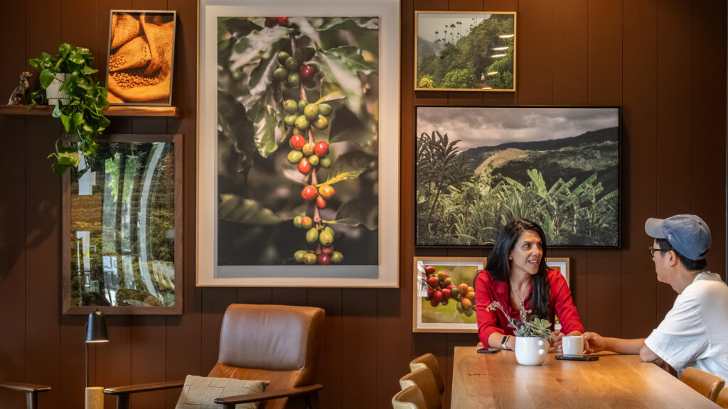 A cozy interior features a woman in a red shirt sitting at a wooden table with a small plant. Behind her, large framed photographs of coffee plants and lush landscapes adorn the walls. A comfortable brown chair is positioned nearby.