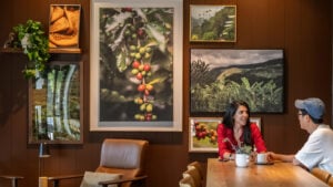 A cozy interior features a woman in a red shirt sitting at a wooden table with a small plant. Behind her, large framed photographs of coffee plants and lush landscapes adorn the walls. A comfortable brown chair is positioned nearby.