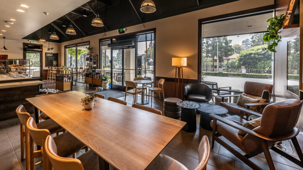 A modern café interior featuring a large wooden table, various seating options, a lamp, and large windows allowing natural light.