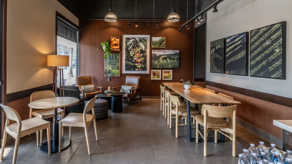 A cozy café interior featuring brown walls, wooden furniture, and various framed nature photographs. There are two armchairs, a small round table, and a long dining table with multiple chairs. A plant adds a touch of greenery.