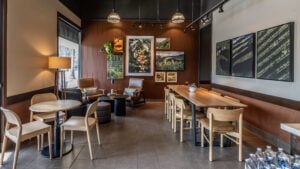 A cozy café interior featuring brown walls, wooden furniture, and various framed nature photographs. There are two armchairs, a small round table, and a long dining table with multiple chairs. A plant adds a touch of greenery.