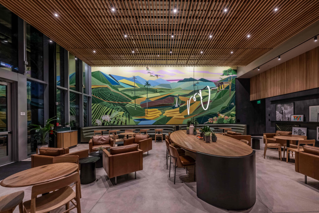 A modern café interior featuring a large geometric mural of green hills and mountains. The space includes leather seating, wooden tables, and plants, creating a cozy atmosphere.