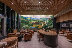 A modern café interior featuring a large geometric mural of green hills and mountains. The space includes leather seating, wooden tables, and plants, creating a cozy atmosphere.