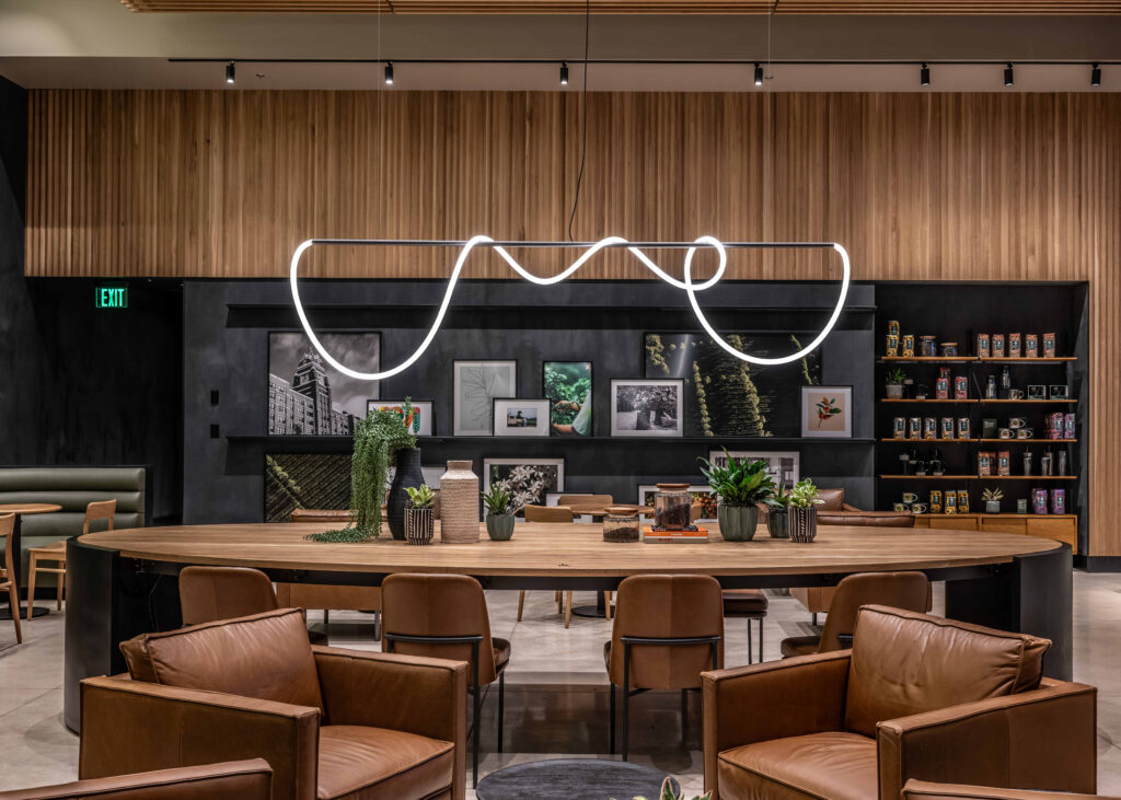 A modern interior featuring a curved, minimalist light fixture above a wooden table. The backdrop includes framed artwork and plants, with leather chairs arranged around the table.