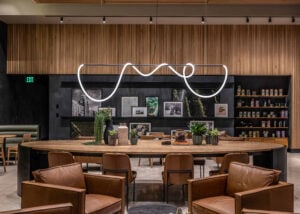 A modern interior featuring a curved, minimalist light fixture above a wooden table. The backdrop includes framed artwork and plants, with leather chairs arranged around the table.