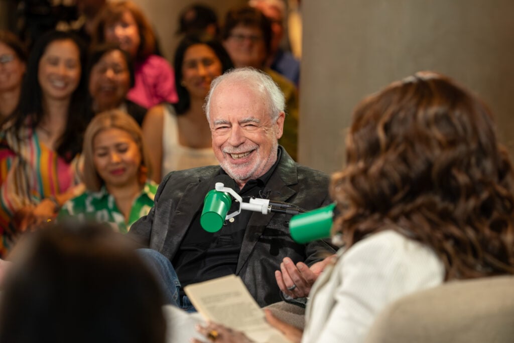 A smiling man with gray hair and a beard is seated, wearing a black jacket. He appears to be engaged in conversation, with a green microphone in front of him. In the background, a diverse audience is visible, showing interest in the discussion.