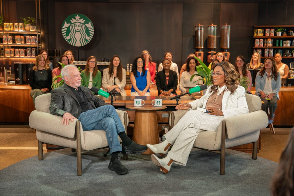 A man and a woman are seated in modern chairs during a discussion. The man wears a black jacket and jeans, while the woman is dressed in a light-colored suit. They are surrounded by an audience of women in a stylish setting with a Starbucks logo in the background. Two mugs with the Starbucks logo are on the table in front of them.