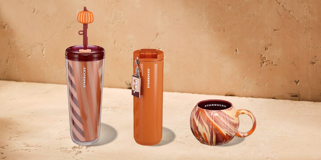 Three Starbucks drinkware items are displayed: a tall tumbler with a pumpkin-shaped lid, a sleek orange travel mug with a tag, and a round marbled mug with a red interior. The background is a textured beige surface.