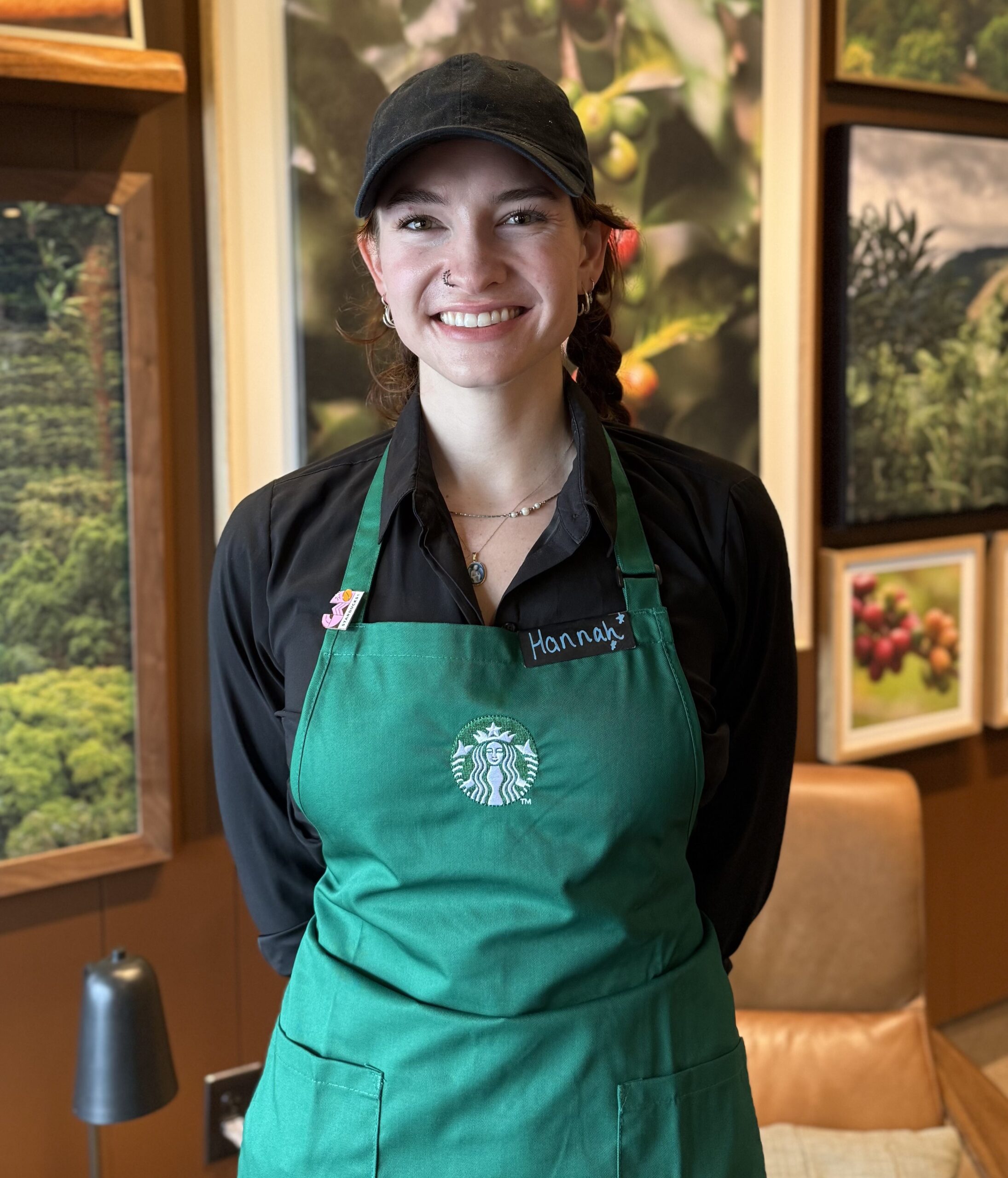 A smiling barista wearing a green Starbucks apron and a black cap stands in front of a wall adorned with coffee-themed artwork.
