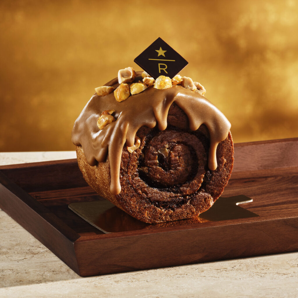A chocolate roll cake topped with a glossy caramel glaze and sprinkled with chopped nuts, presented on a wooden tray.