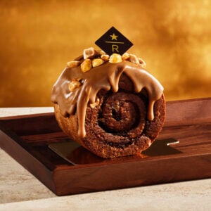 A chocolate roll cake topped with a glossy caramel glaze and sprinkled with chopped nuts, presented on a wooden tray.