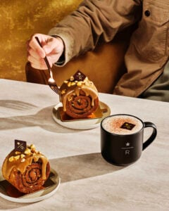 A hand holds a fork poised to cut into a cinnamon roll topped with caramel and nuts, alongside a black mug of frothy coffee.
