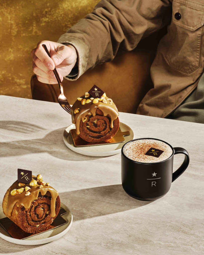 A hand holds a fork poised to cut into a cinnamon roll topped with caramel and nuts, alongside a black mug of frothy coffee.