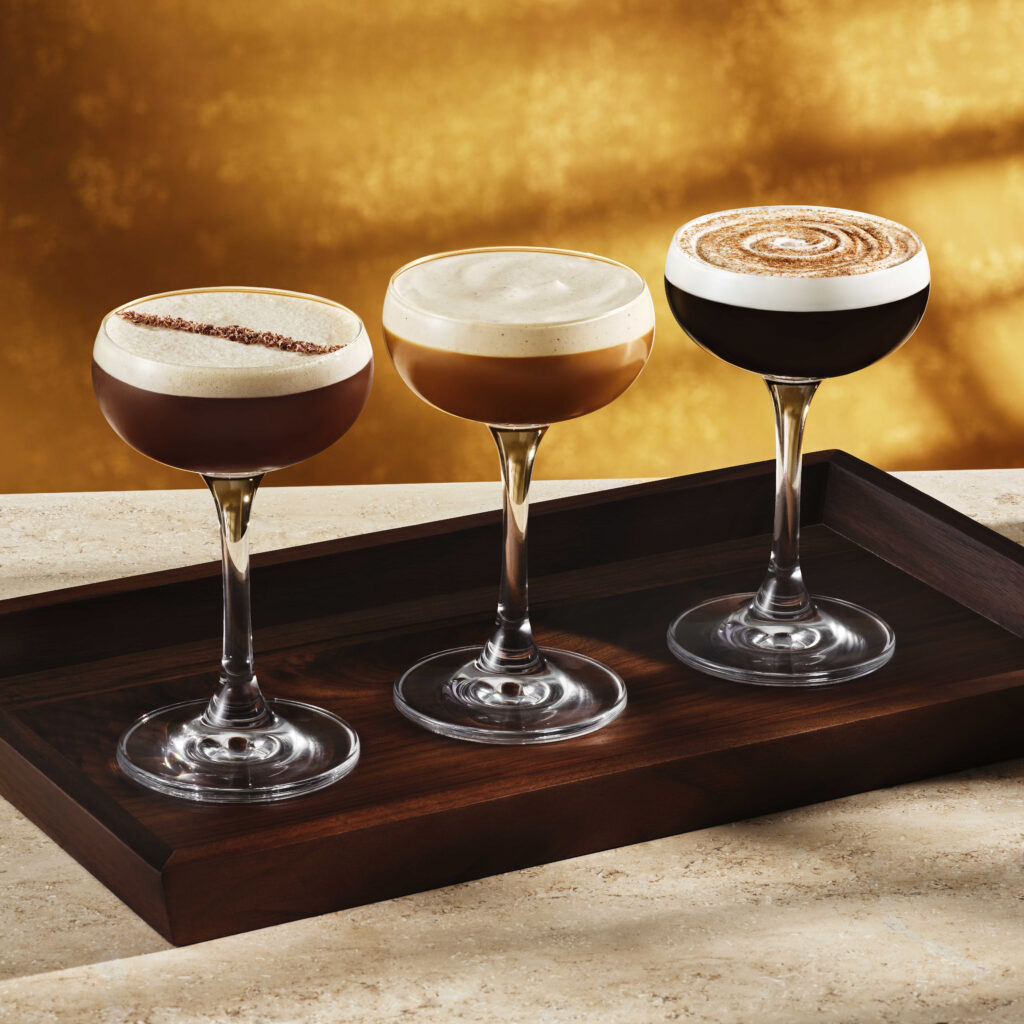Three elegant cocktail glasses are displayed on a wooden tray. Each glass contains a different layered drink, with creamy froth on top. The background features a warm golden hue, enhancing the sophisticated presentation.