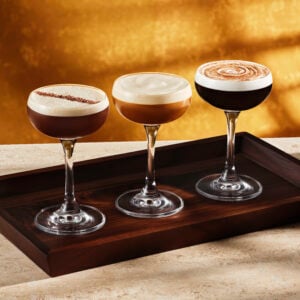 Three elegant cocktail glasses are displayed on a wooden tray. Each glass contains a different layered drink, with creamy froth on top. The background features a warm golden hue, enhancing the sophisticated presentation.