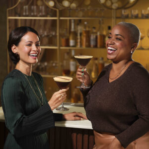 Two women are joyfully toasting with cocktails at a stylish bar. One wears a dark green pleated dress, while the other is in a brown sweater. The background features shelves of bottles, creating a warm ambiance.
