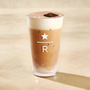 A tall glass of iced coffee with a creamy layer on top, featuring a chocolate piece and a logo with a star and "R.