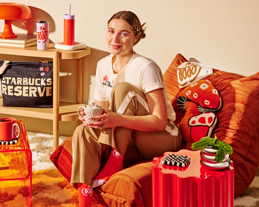 A person sits on a vibrant orange cushion, holding a mug with whipped cream. They wear a light shirt and beige pants with stripes, complemented by red socks. The background features colorful decor, including a patterned pillow, a small red table, and a plant.