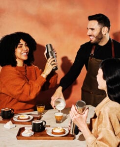 A group of three people enjoy coffee and pastries at a table. One person shakes a cocktail shaker, while another pours milk into a glass. Plates with desserts and mugs are arranged on a wooden tray. The background features a warm, textured wall.