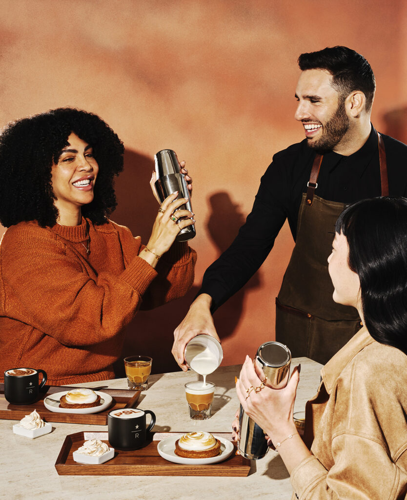 A group of three people enjoy coffee and pastries at a table. One person shakes a cocktail shaker, while another pours milk into a glass. Plates with desserts and mugs are arranged on a wooden tray. The background features a warm, textured wall.