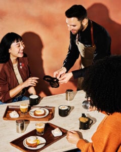 A barista serves a woman at a table with coffee drinks and desserts. Two mugs are present, one with "PSL" and another with "R." The setting has warm tones and a cozy atmosphere.